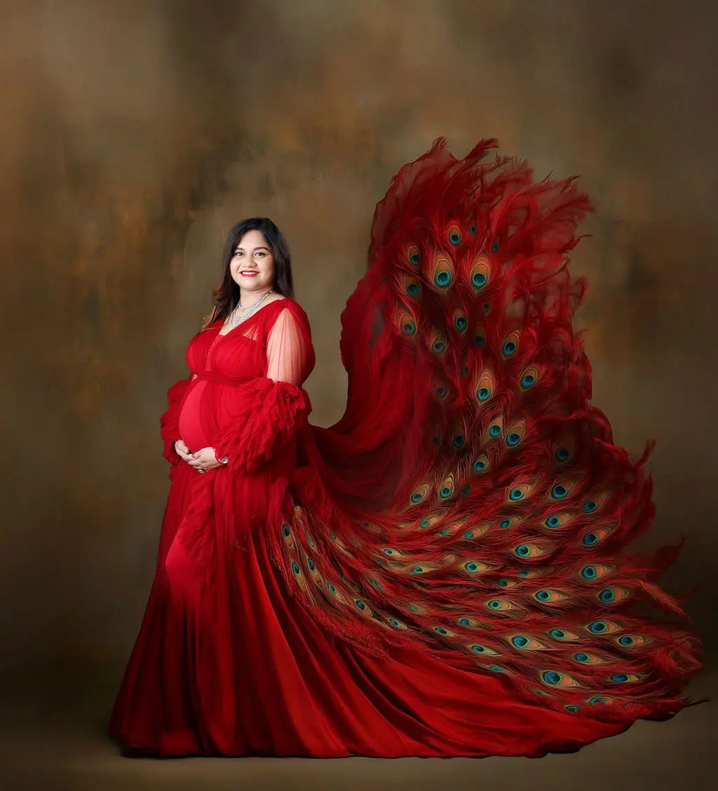 Smiling expectant mother in a red tulle gown with a flowing peacock feather train on a painted backdrop, maternity photoshoot at WonderBump Studio, Bengaluru