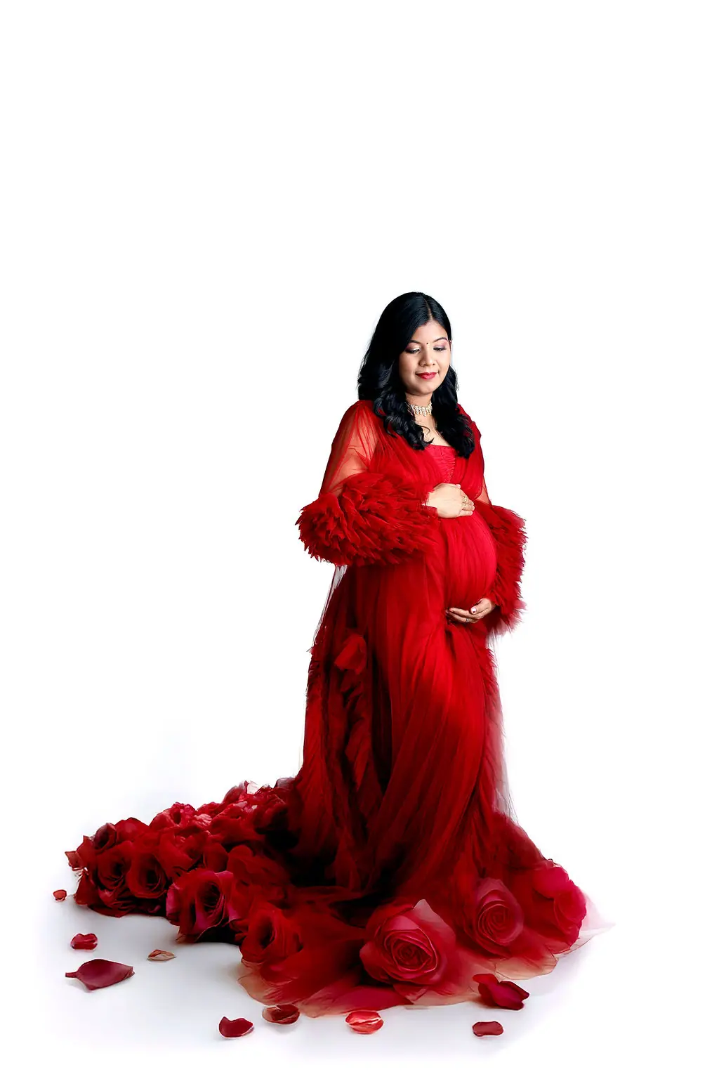 Expectant mother cradling her bump in a red tulle gown with a rose train and scattered petals on white, maternity photoshoot at WonderBump Studio, Bengaluru