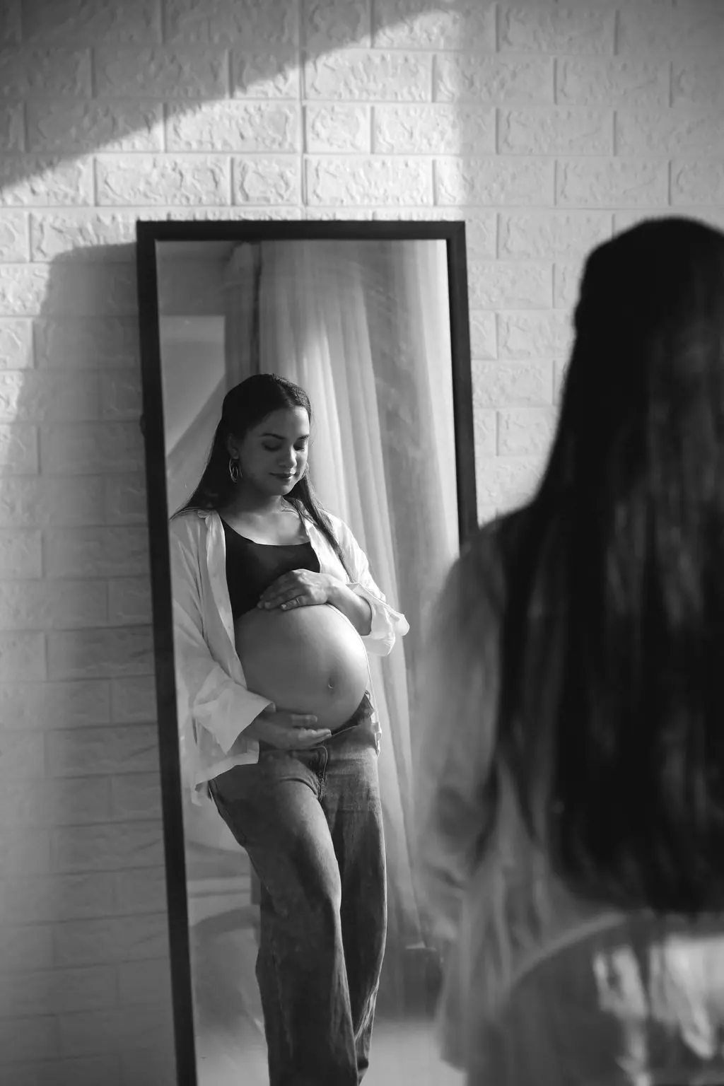Expectant mother reflected in a tall mirror, cradling her bump in an open white shirt and jeans, maternity photoshoot at WonderBump Studio, Bengaluru