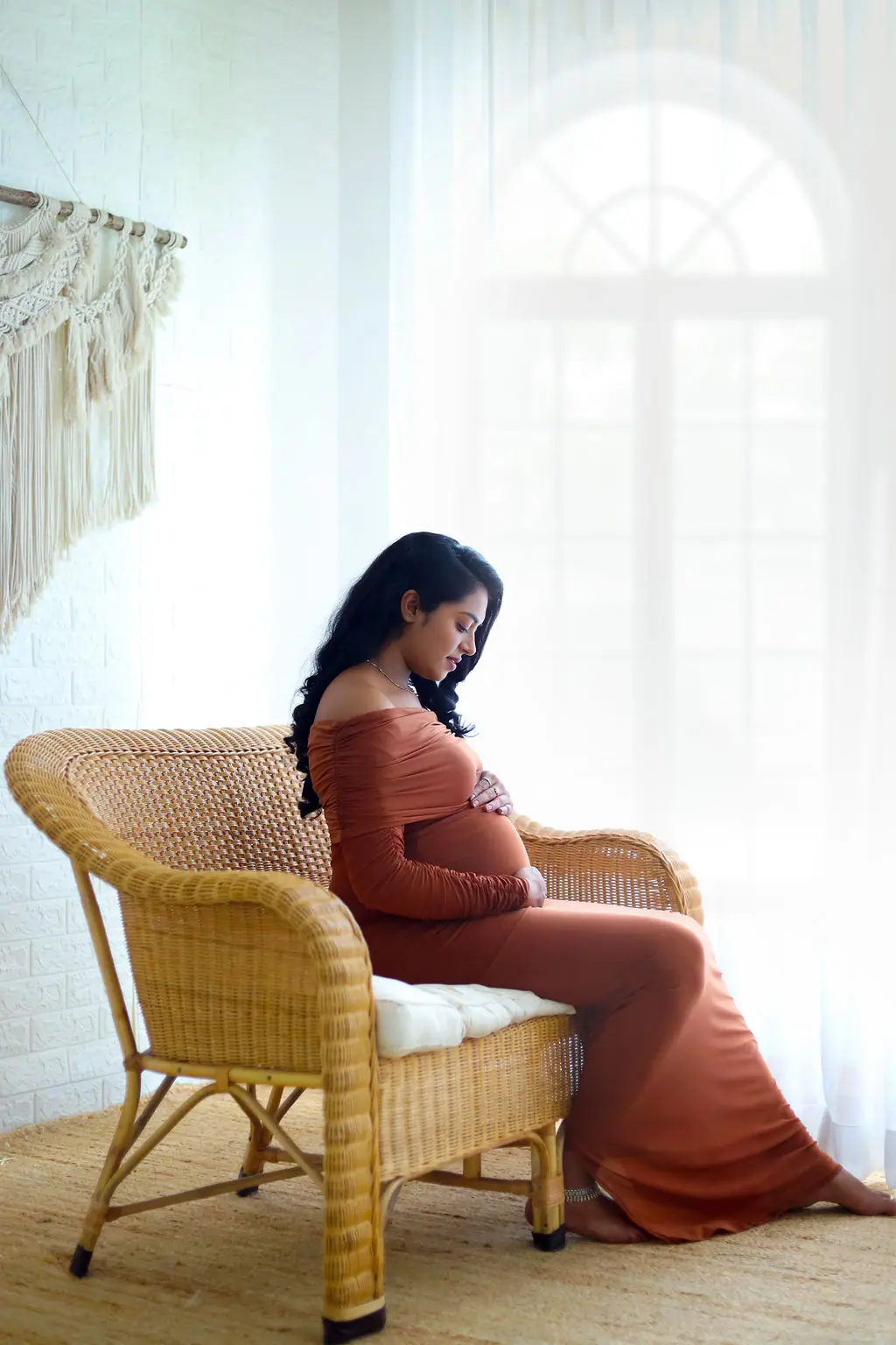Expectant mother seated on a wicker armchair in a rust off-shoulder gown, maternity photoshoot at WonderBump Studio, Bengaluru