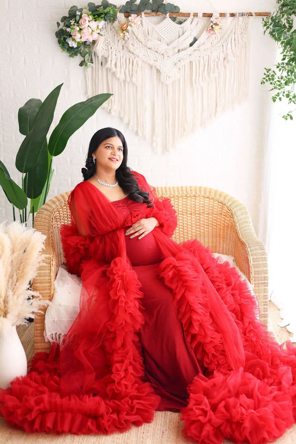 Expectant mother seated on a wicker sofa in a red ruffled tulle robe, macrame and greenery, maternity photoshoot at WonderBump Studio, Bengaluru