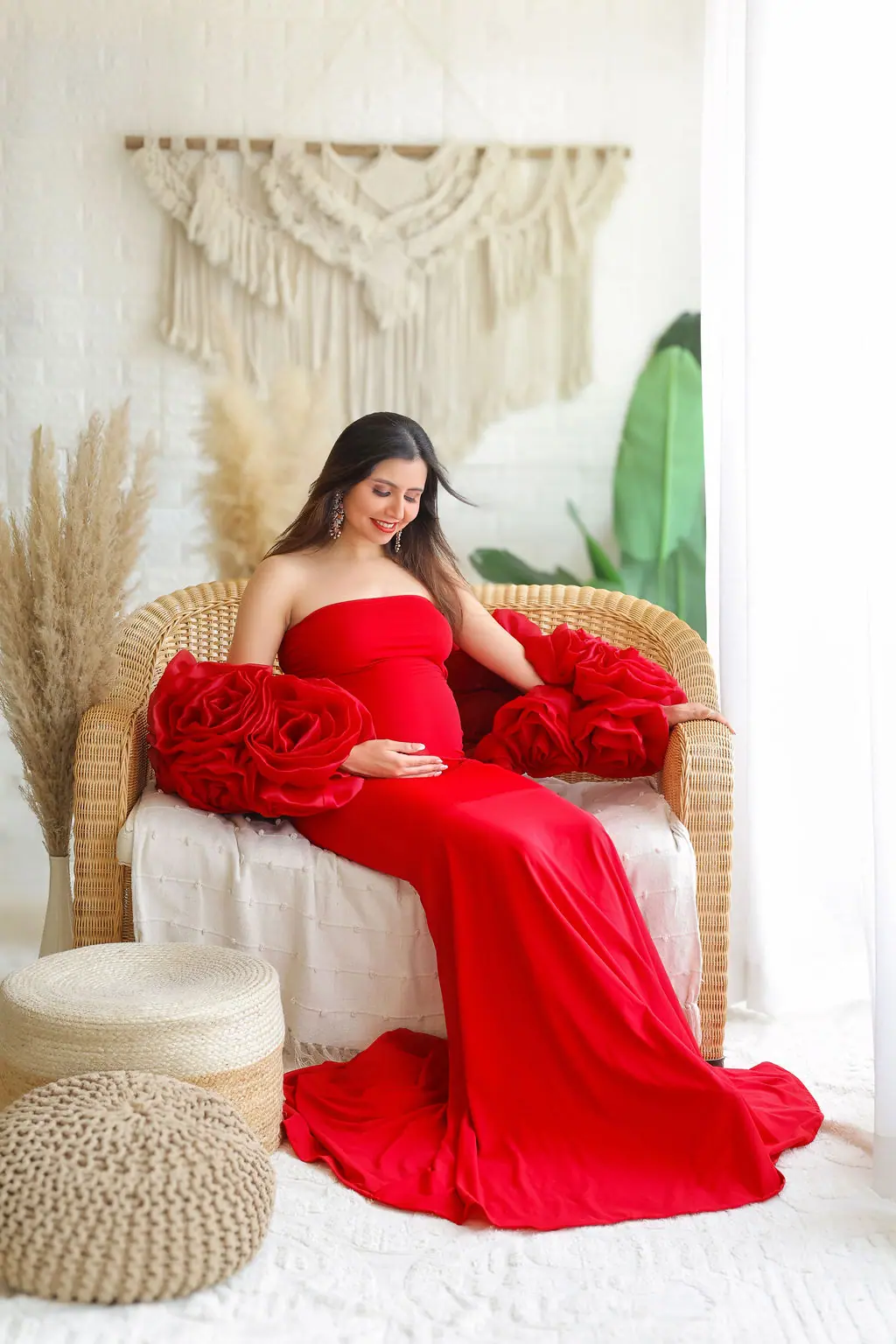Expectant mother seated on a wicker sofa in a red strapless gown with rose sleeves, maternity photoshoot at WonderBump Studio, Bengaluru