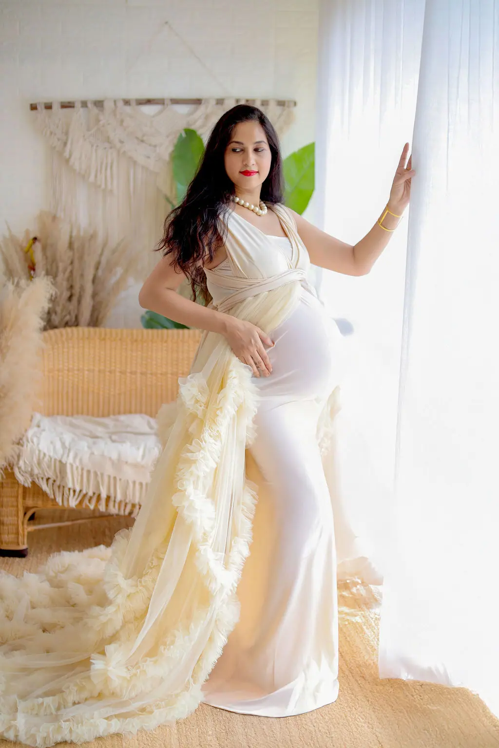 Expectant mother by a sheer curtain in a cream ruffled tulle gown in a boho set, maternity photoshoot at WonderBump Studio, Bengaluru