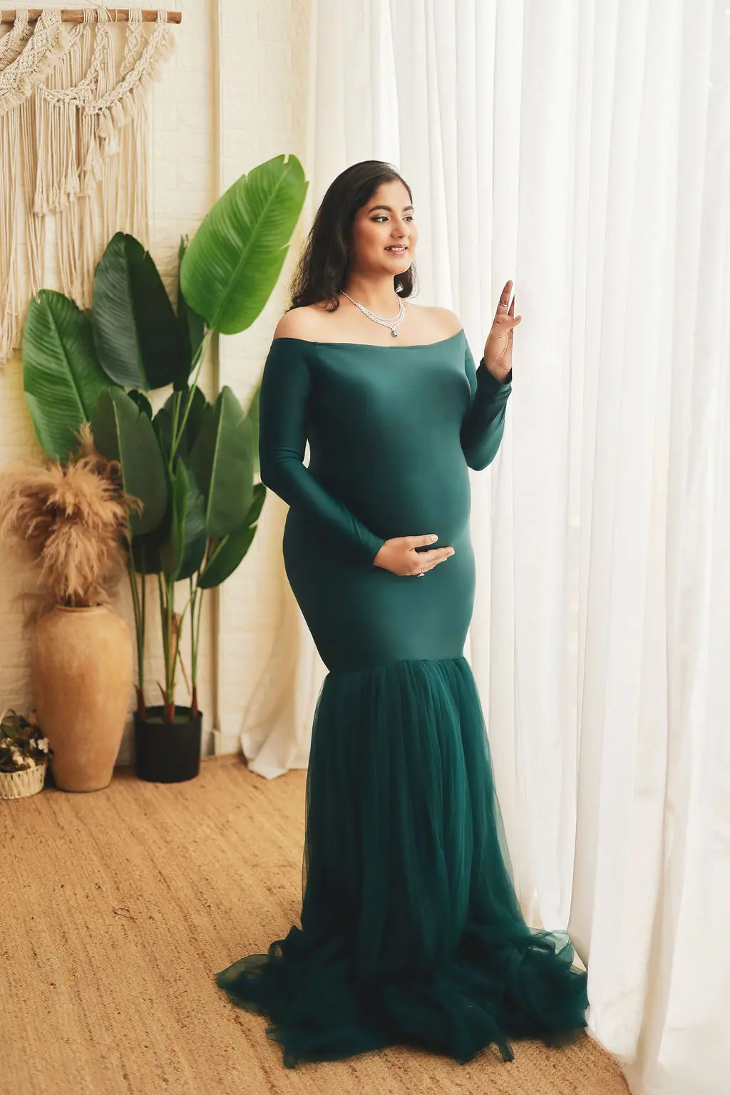 Expectant mother by sheer curtains in an off-shoulder emerald mermaid gown with leafy plants and macrame, maternity photoshoot at WonderBump Studio, Bengaluru