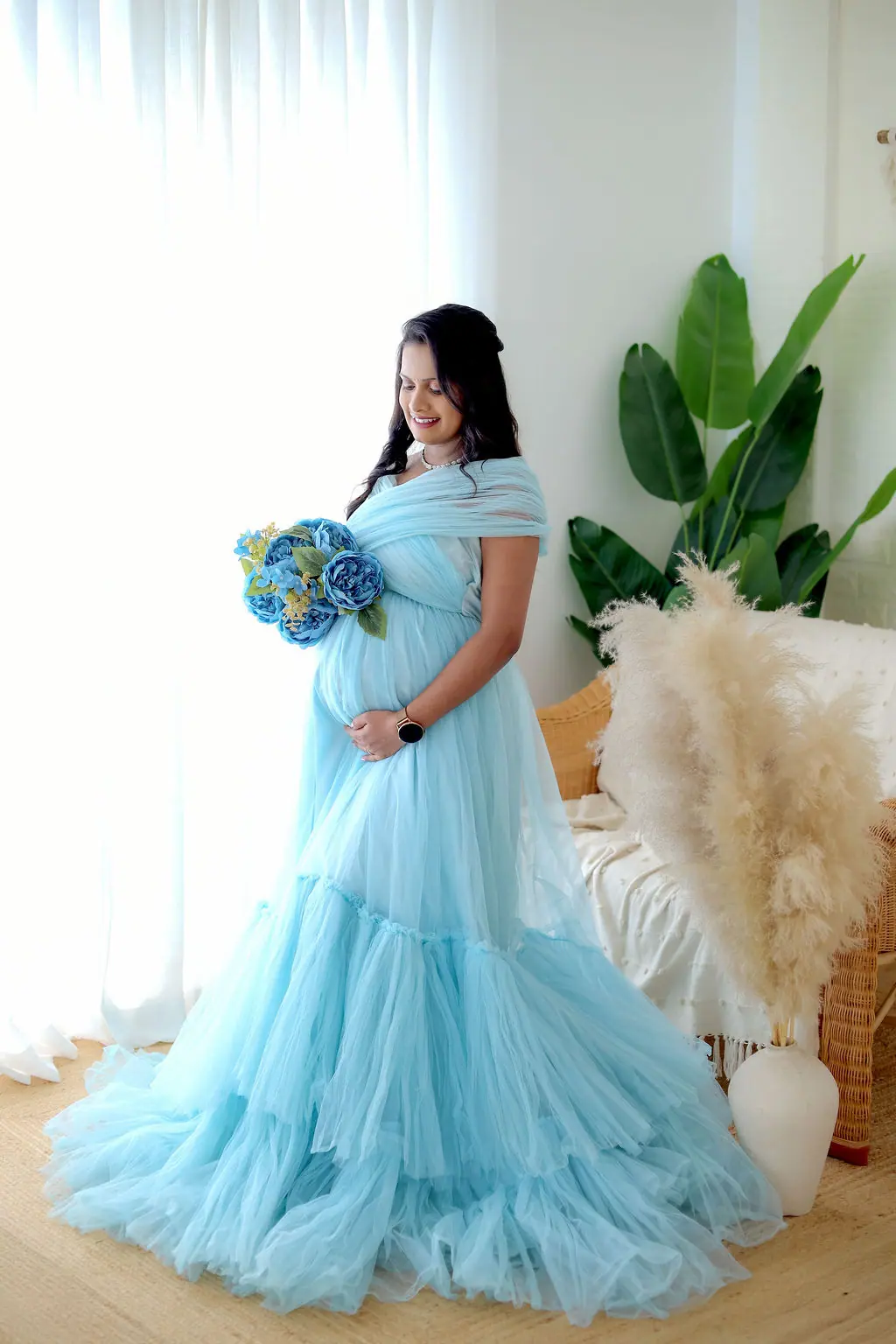 Expectant mother holding blue flowers in a sky blue tulle gown beside pampas grass, maternity photoshoot at WonderBump Studio, Bengaluru