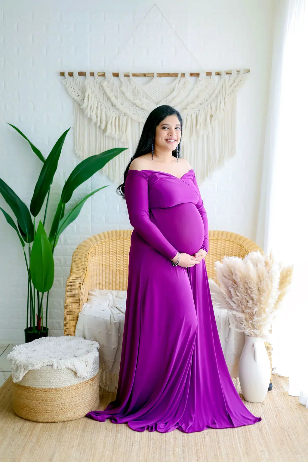 Smiling expectant mother in a magenta off-shoulder gown beside a wicker chair and pampas grass, maternity photoshoot at WonderBump Studio, Bengaluru