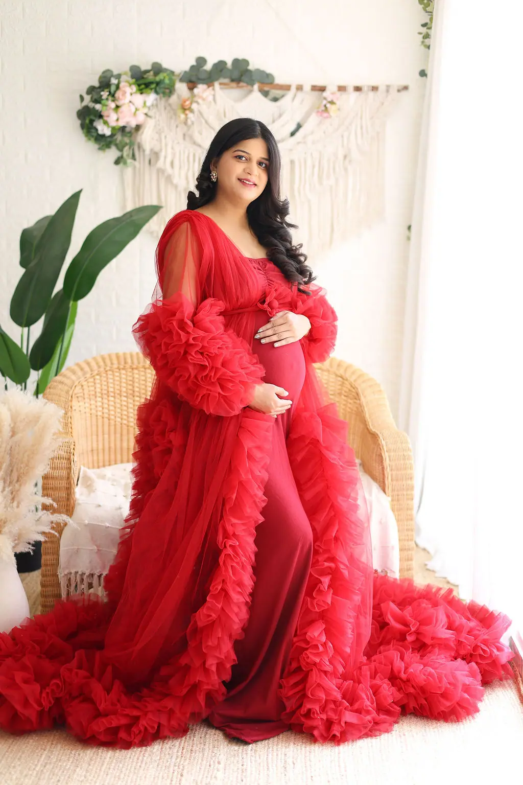 Smiling expectant mother in a red ruffled tulle robe before a wicker chair and macrame wall, maternity photoshoot at WonderBump Studio, Bengaluru