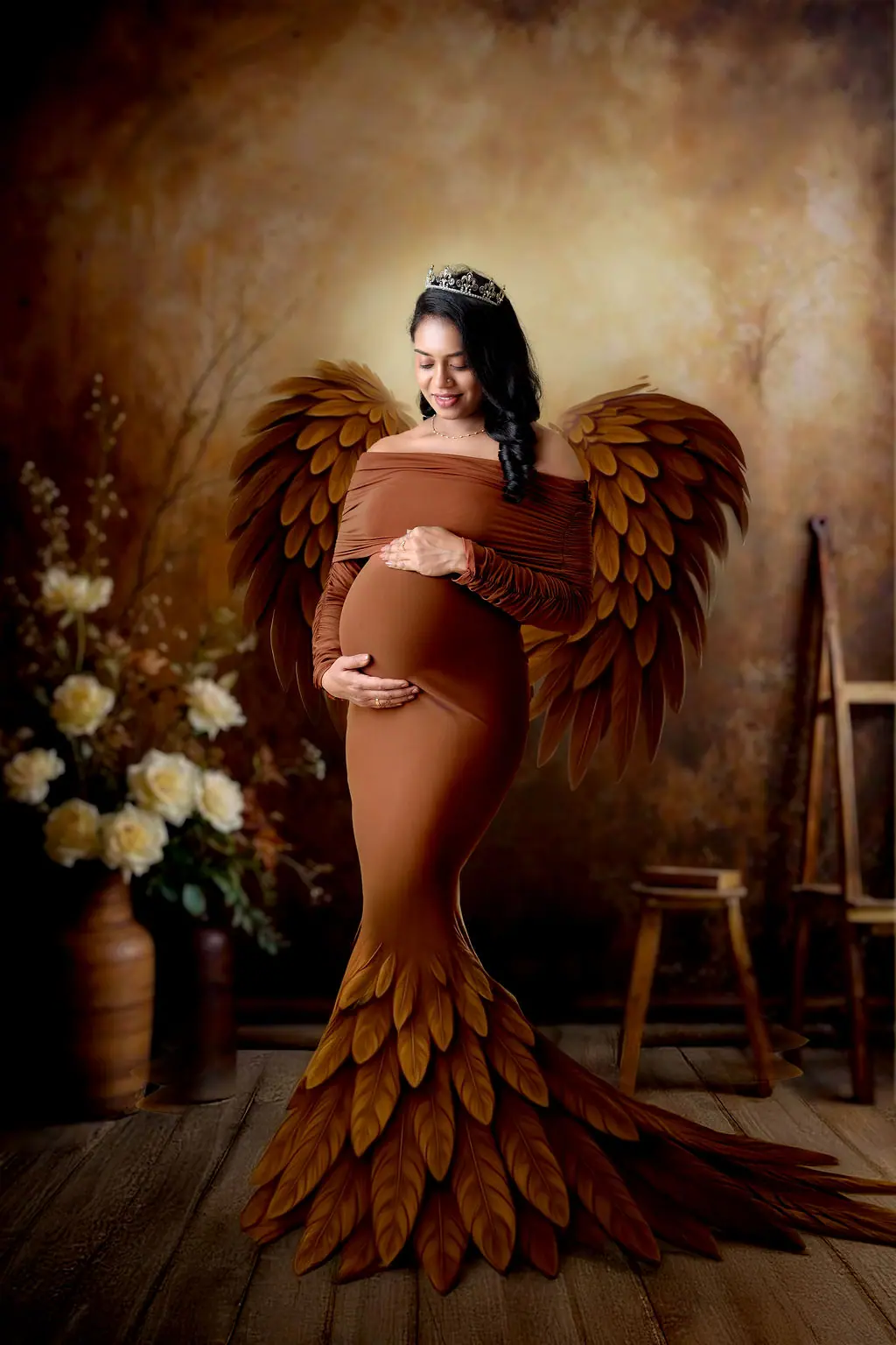 Expectant mother in a tiara and brown gown with feathered wings on a painted backdrop, maternity photoshoot at WonderBump Studio, Bengaluru