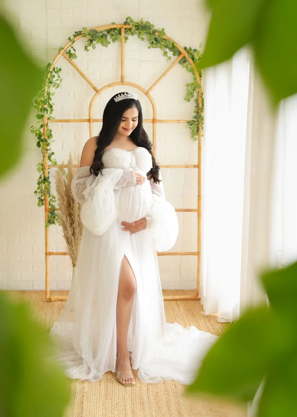 Expectant mother in a tiara and white pearl tulle gown by a rattan arch with vines, maternity photoshoot at WonderBump Studio, Bengaluru