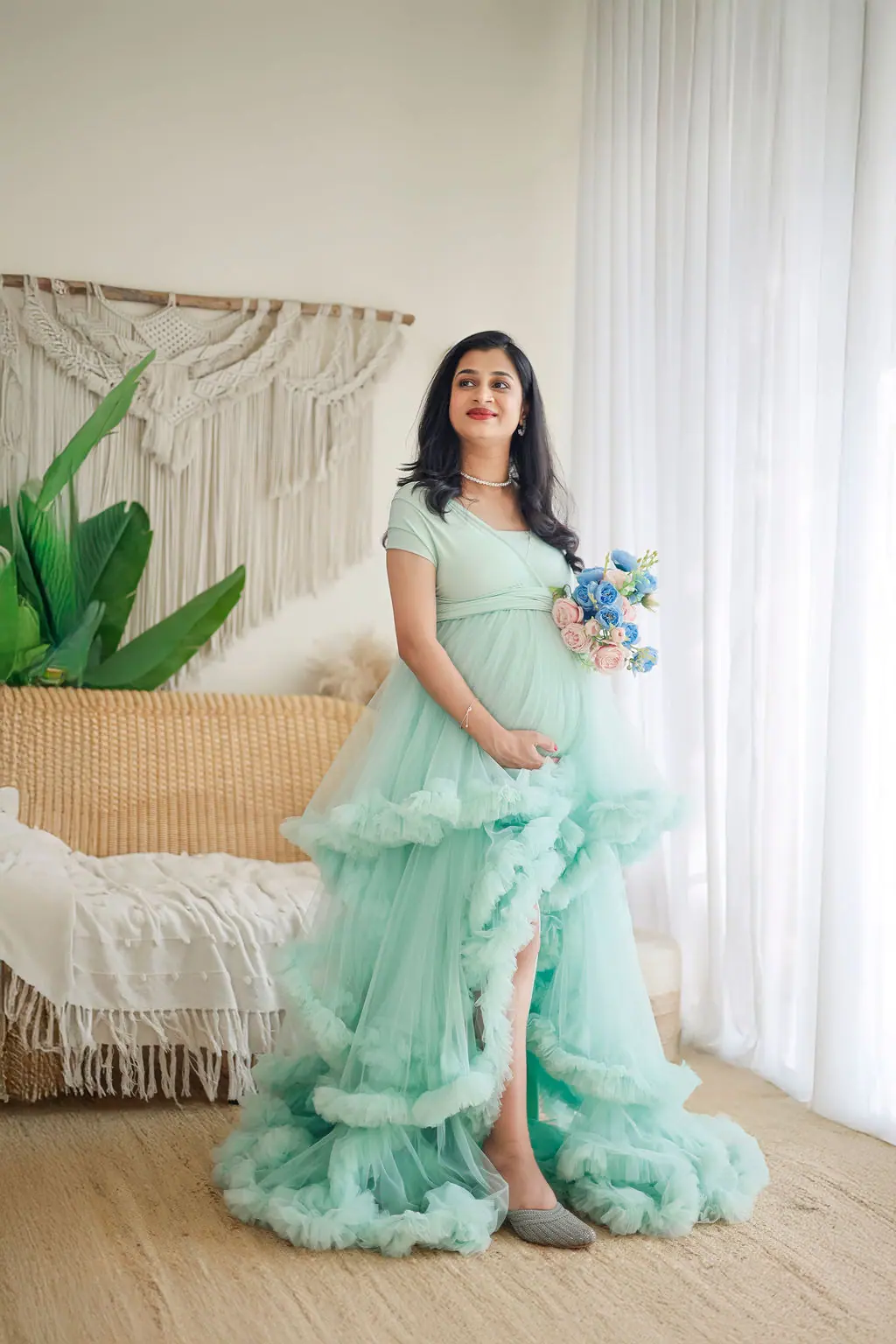Expectant mother with a blue and pink bouquet in a tiered mint tulle gown on a boho macrame set, maternity photoshoot at WonderBump Studio, Bengaluru