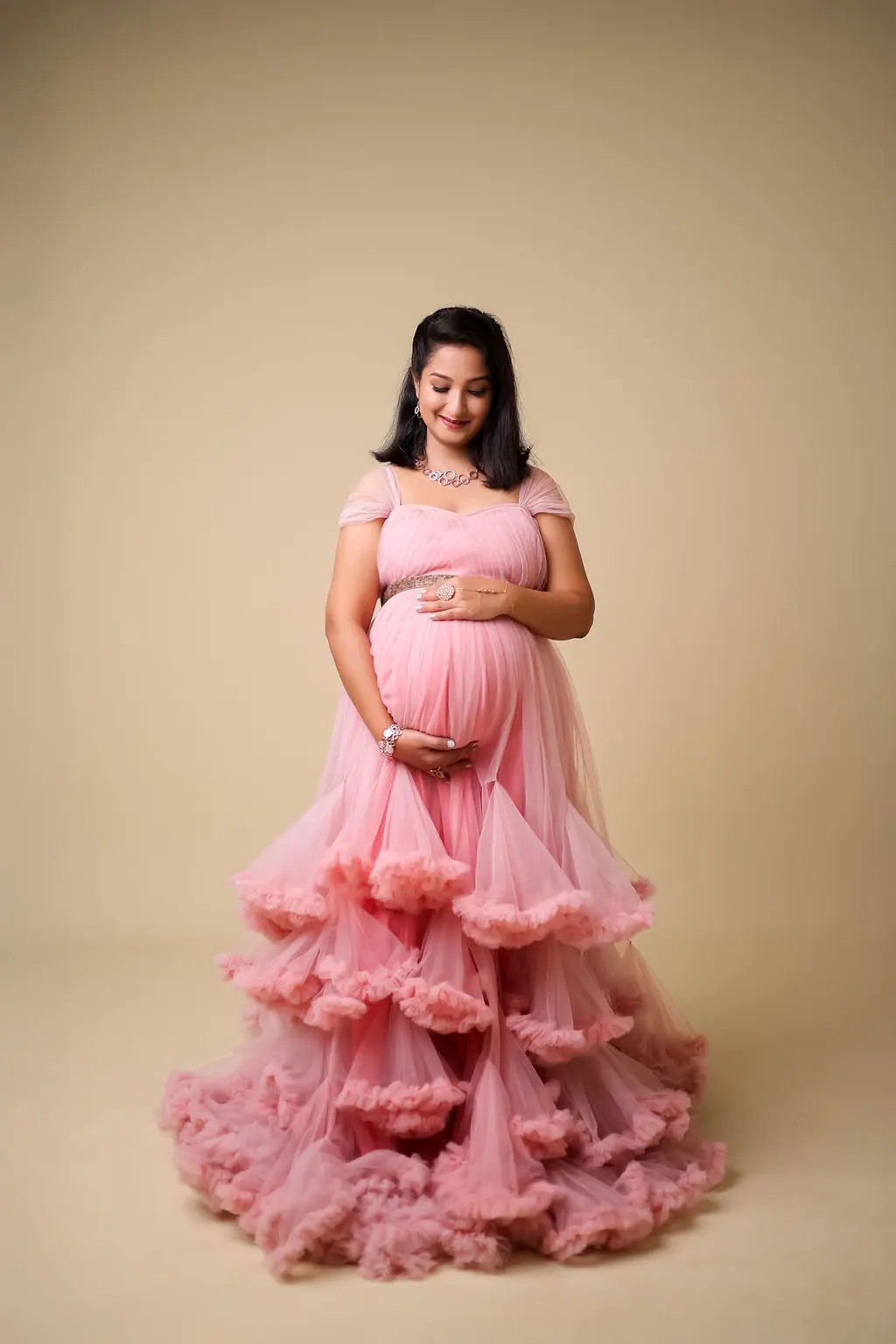 Expectant mother in a tiered pink ruffled tulle gown gazing down at her bump, beige backdrop, maternity photoshoot at WonderBump Studio, Bengaluru
