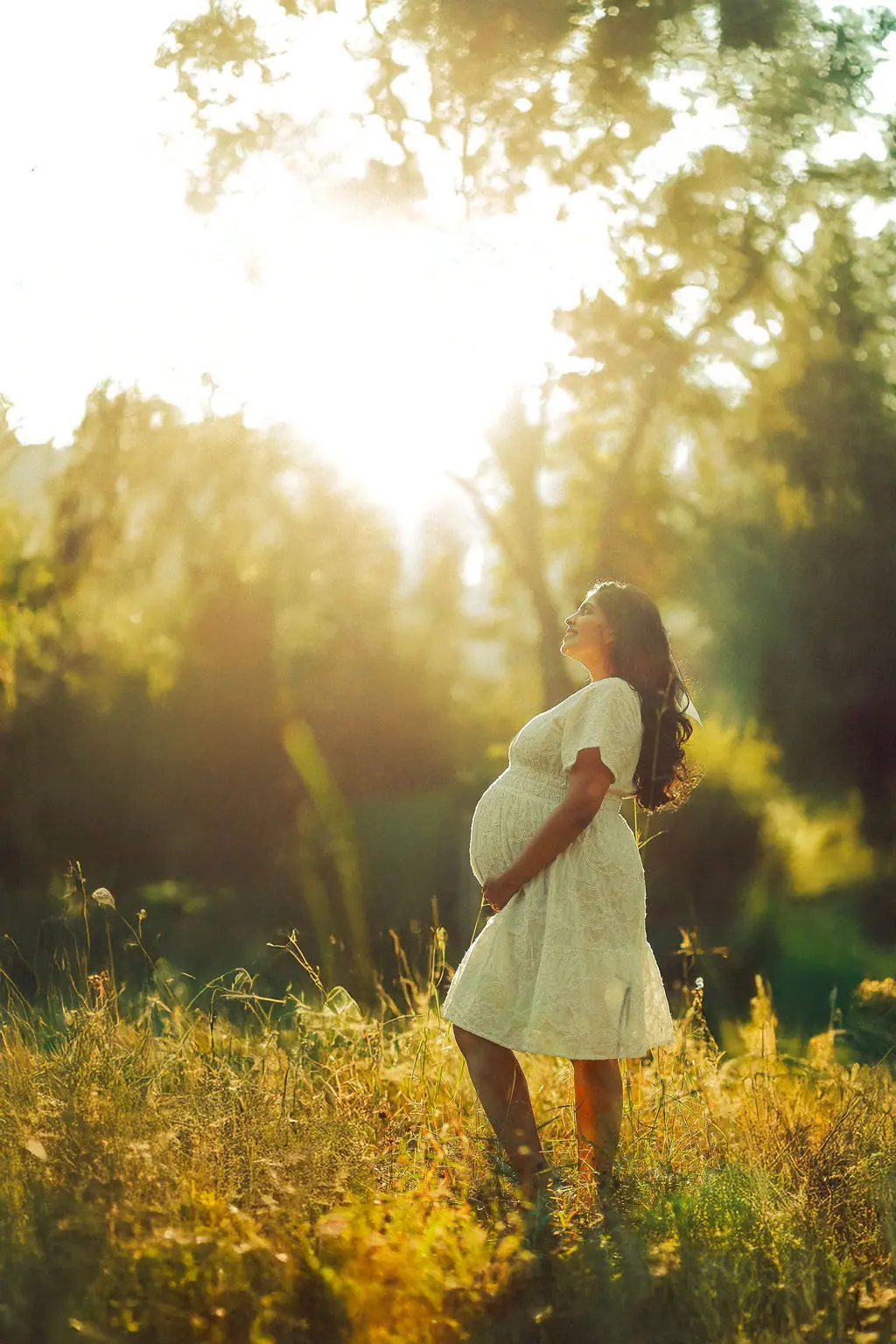 Expectant mother in a white dress cradling her bump in tall golden grass under glowing sunlight, maternity photoshoot by WonderBump Studio, Bengaluru