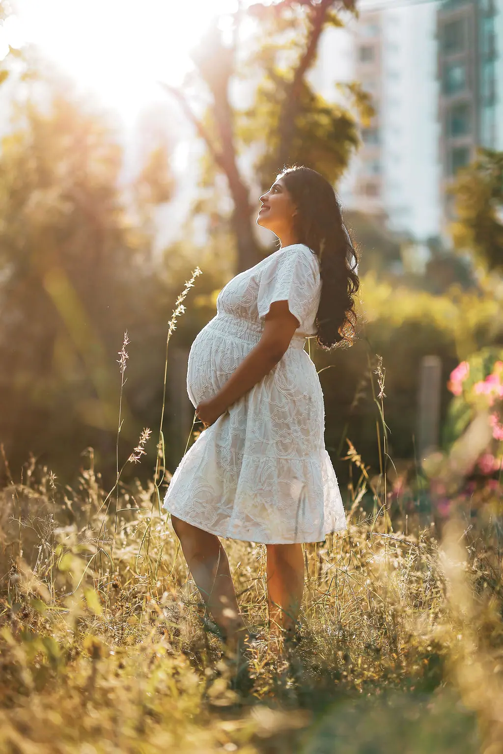 Expectant mother in profile in a white embroidered dress, smiling skyward among tall grass at golden hour, maternity photoshoot by WonderBump Studio, Bengaluru