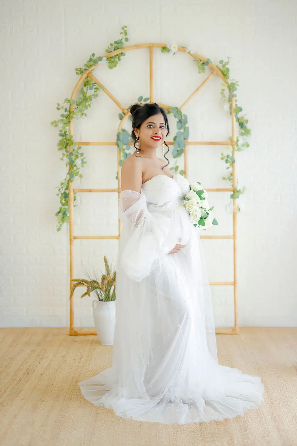 Smiling expectant mother in a white pearl tulle gown holding white roses before a rattan arch with vines, maternity photoshoot at WonderBump Studio, Bengaluru