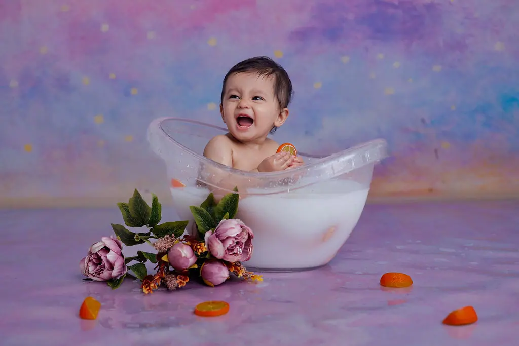 Laughing baby in a clear milk bath tub holding an orange slice, with peonies and a painted pastel backdrop, baby photoshoot at WonderBump Studio, Bengaluru