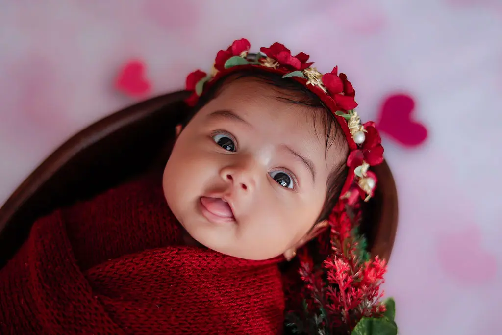Curious baby in a red knit wrap and red flower crown lying in a wooden bowl with paper hearts, baby photoshoot at WonderBump Studio, Bengaluru