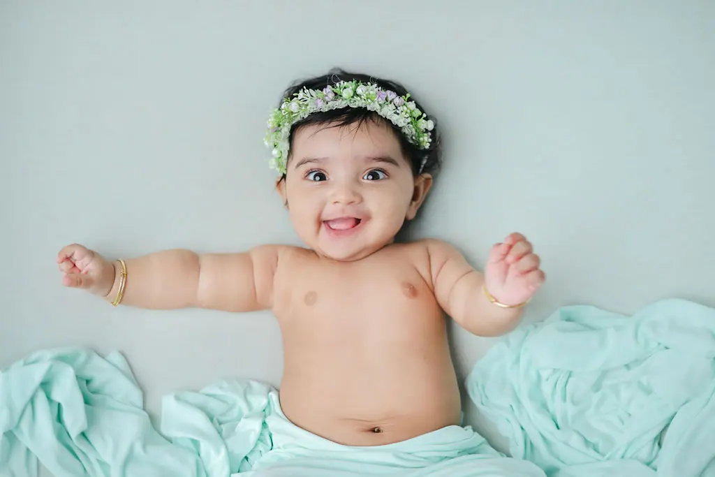 Smiling baby lying on back with arms out, wearing a floral crown and bangles on a mint wrap, baby photoshoot at WonderBump Studio, Bengaluru