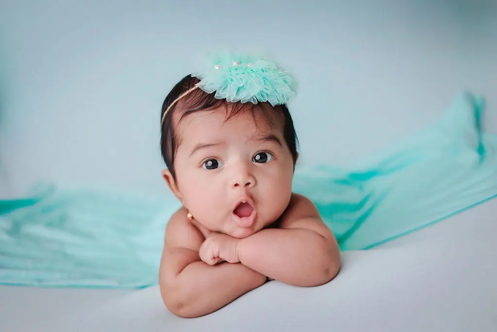 Surprised baby lying on tummy with arms folded, wearing a mint tulle headband on a pale blue set, baby photoshoot at WonderBump Studio, Bengaluru