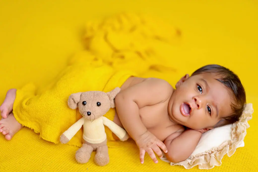 Wide-eyed baby lying on a frilled pillow under a yellow knit wrap beside a plush mouse toy, baby photoshoot at WonderBump Studio, Bengaluru