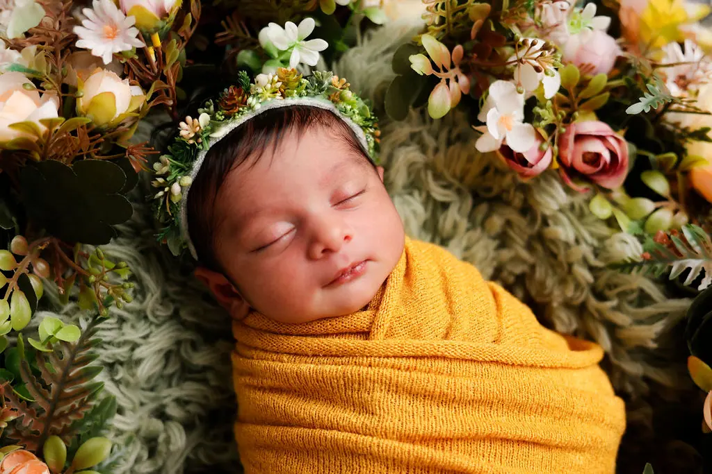Close-up of a sleeping newborn in a flower crown and mustard wrap on green faux fur among blooms, newborn photoshoot at WonderBump Studio, Bengaluru