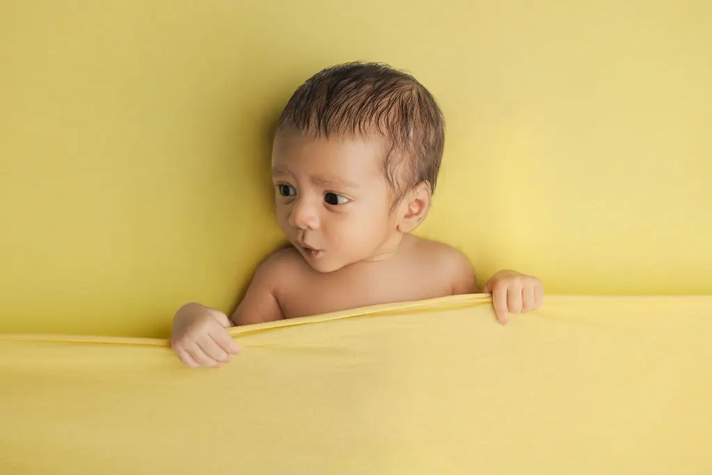 Curious newborn peeking over a yellow blanket, tiny hands gripping its edge, newborn photoshoot at WonderBump Studio, Bengaluru