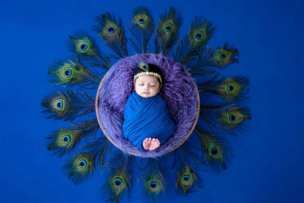 Sleeping newborn in a blue wrap and gold crown ringed by peacock feathers on blue, newborn photoshoot at WonderBump Studio, Bengaluru