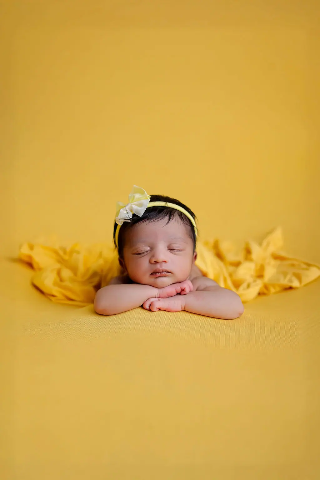 Sleeping newborn resting chin on folded hands, wearing a bow headband on a mustard yellow backdrop, newborn photoshoot at WonderBump Studio, Bengaluru