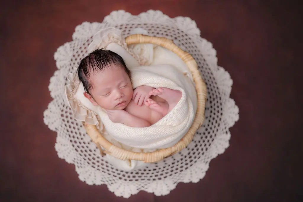 Sleeping newborn curled in a cream knit wrap inside a cane basket on a lace doily, newborn photoshoot at WonderBump Studio, Bengaluru