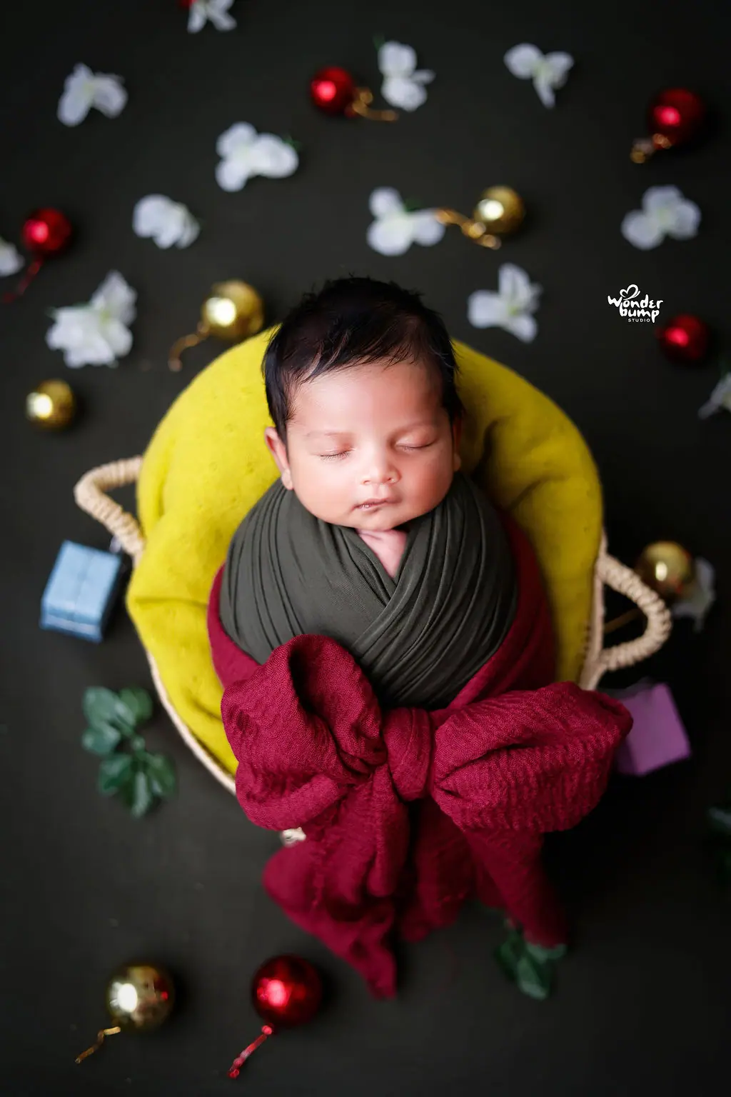 Sleeping newborn in a green wrap and red bow in a basket among festive baubles, newborn photoshoot at WonderBump Studio, Bengaluru