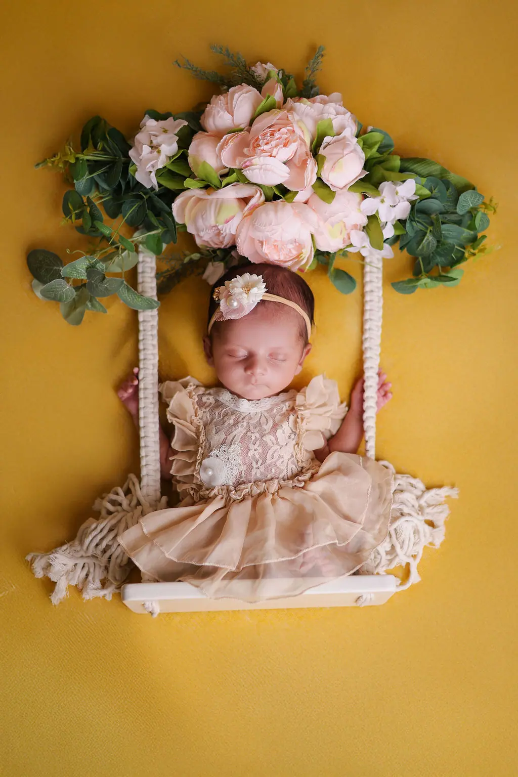 Sleeping newborn in a lace tulle dress on a swing under blush peonies on yellow, newborn photoshoot at WonderBump Studio, Bengaluru