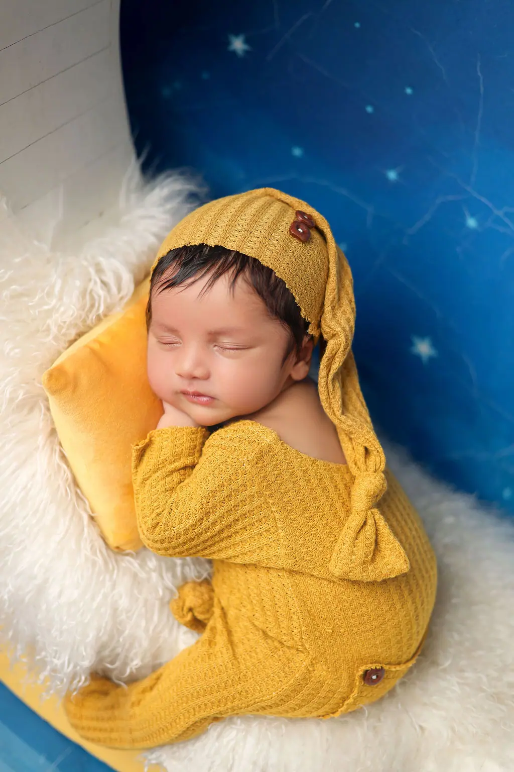 Sleeping newborn in a mustard knit romper and sleepy hat on a moon prop with a painted starry backdrop, newborn photoshoot at WonderBump Studio, Bengaluru
