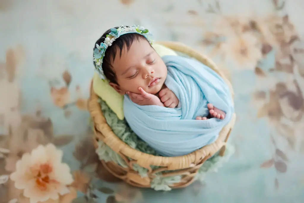 Sleeping newborn in a pale blue wrap and floral headband curled in a cane basket on a floral backdrop, newborn photoshoot at WonderBump Studio, Bengaluru