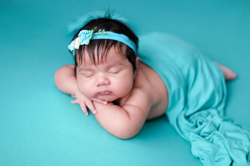 Sleeping newborn resting chin on arms in a teal wrap and flower headband on teal, newborn photoshoot at WonderBump Studio, Bengaluru