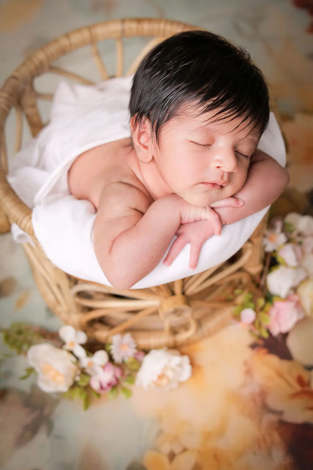 Sleeping newborn resting chin on hands in a rattan chair among soft flowers, newborn photoshoot at WonderBump Studio, Bengaluru
