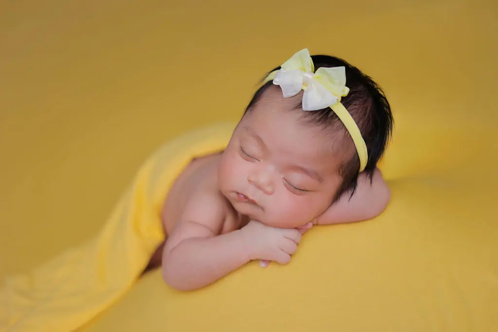 Sleeping newborn resting on folded arms in a yellow wrap and bow headband, newborn photoshoot at WonderBump Studio, Bengaluru
