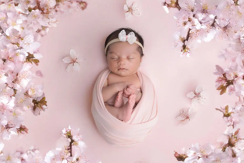 Sleeping newborn swaddled in a blush wrap with a lace bow headband, framed by pink blossoms, newborn photoshoot at WonderBump Studio, Bengaluru