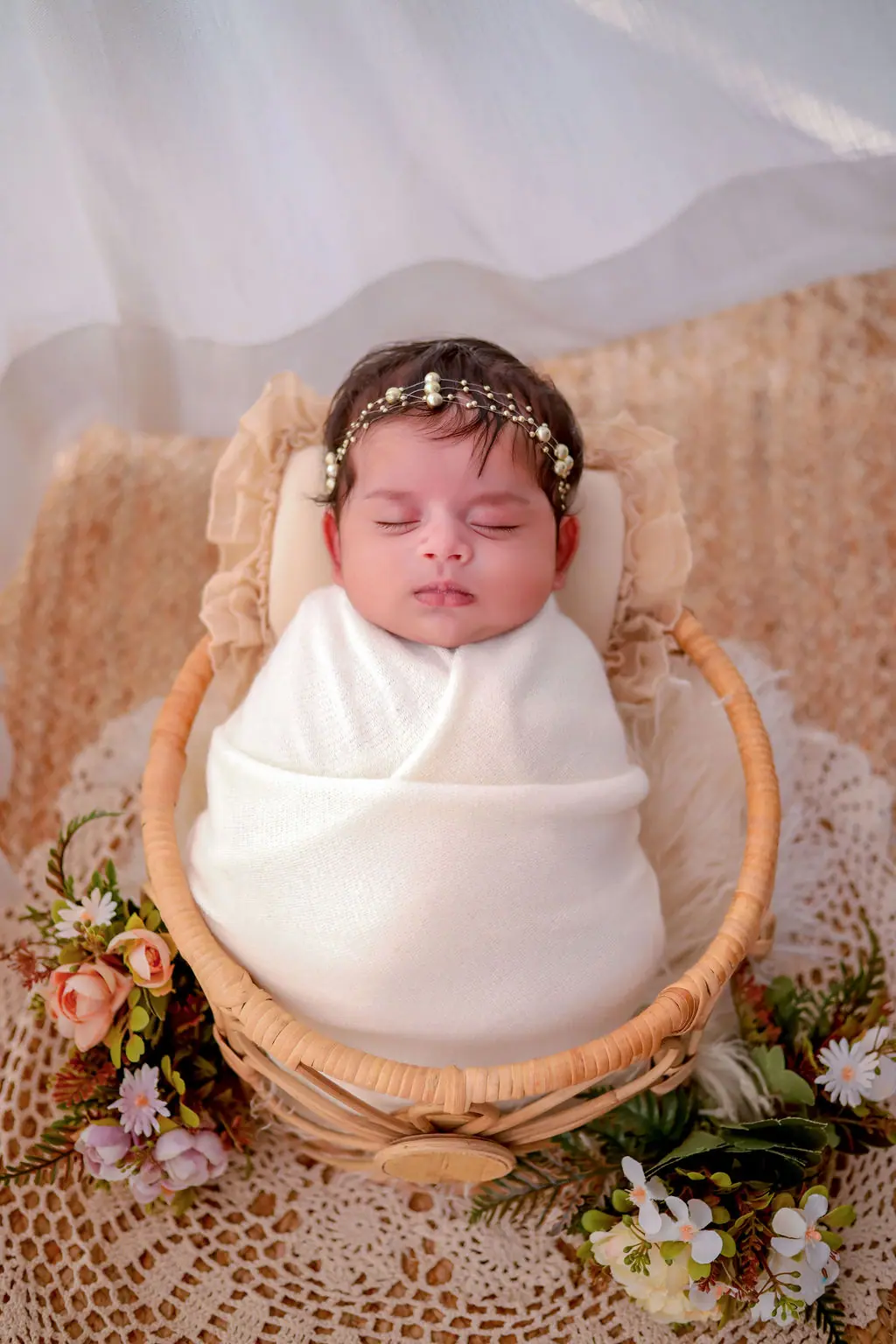 Sleeping newborn swaddled in white with a pearl headpiece in a cane basket, flowers and sheer drapes, newborn photoshoot at WonderBump Studio, Bengaluru