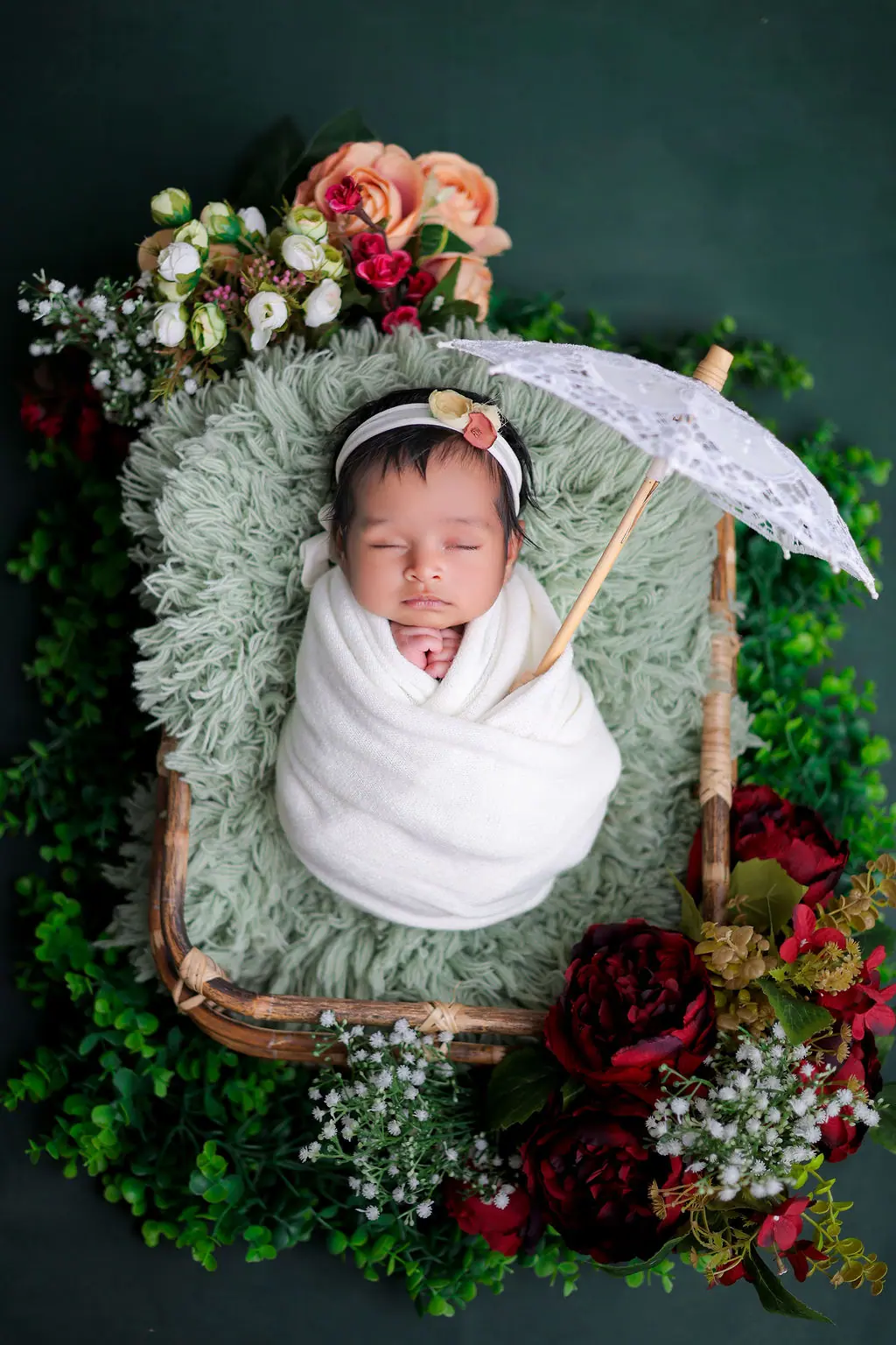 Sleeping newborn in a white wrap in a basket with a lace parasol and roses on green, newborn photoshoot at WonderBump Studio, Bengaluru