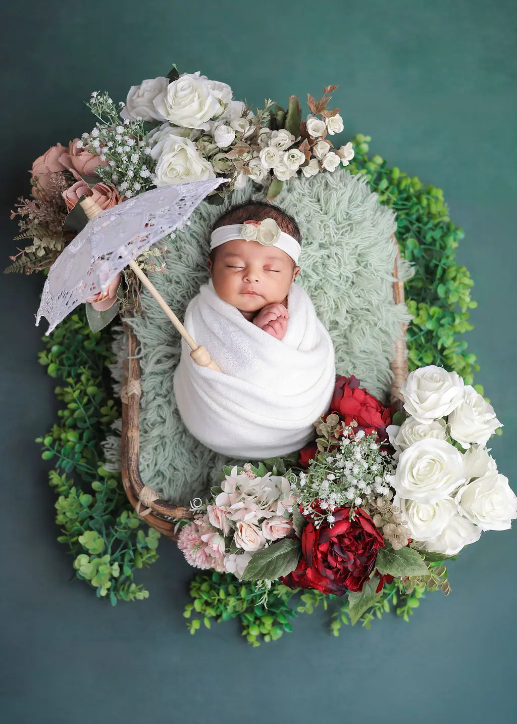 Sleeping newborn in a white wrap in a basket with a lace parasol and roses on teal, newborn photoshoot at WonderBump Studio, Bengaluru