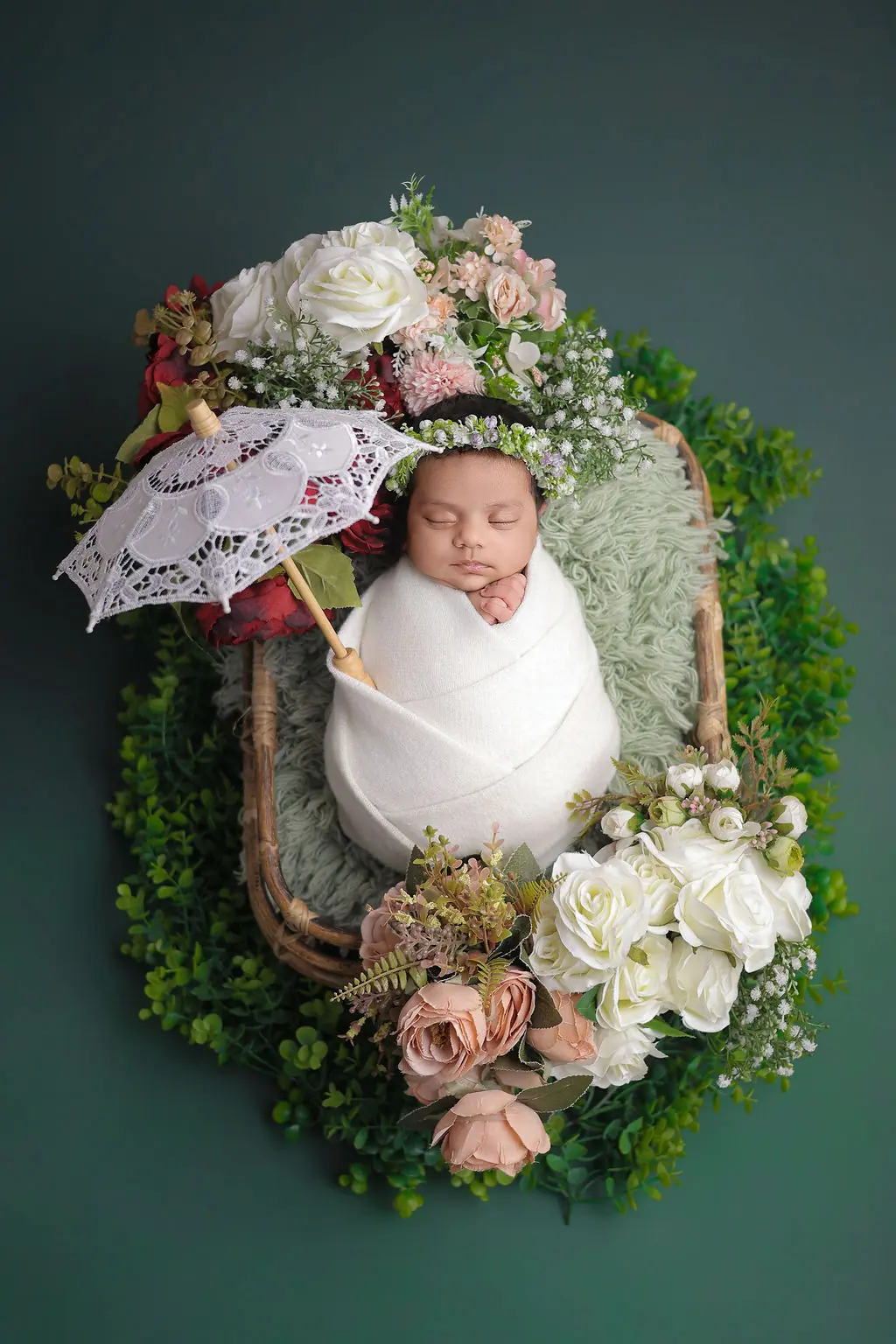Sleeping newborn in a white wrap and floral crown inside a rattan basket with roses and a lace parasol, newborn photoshoot at WonderBump Studio, Bengaluru