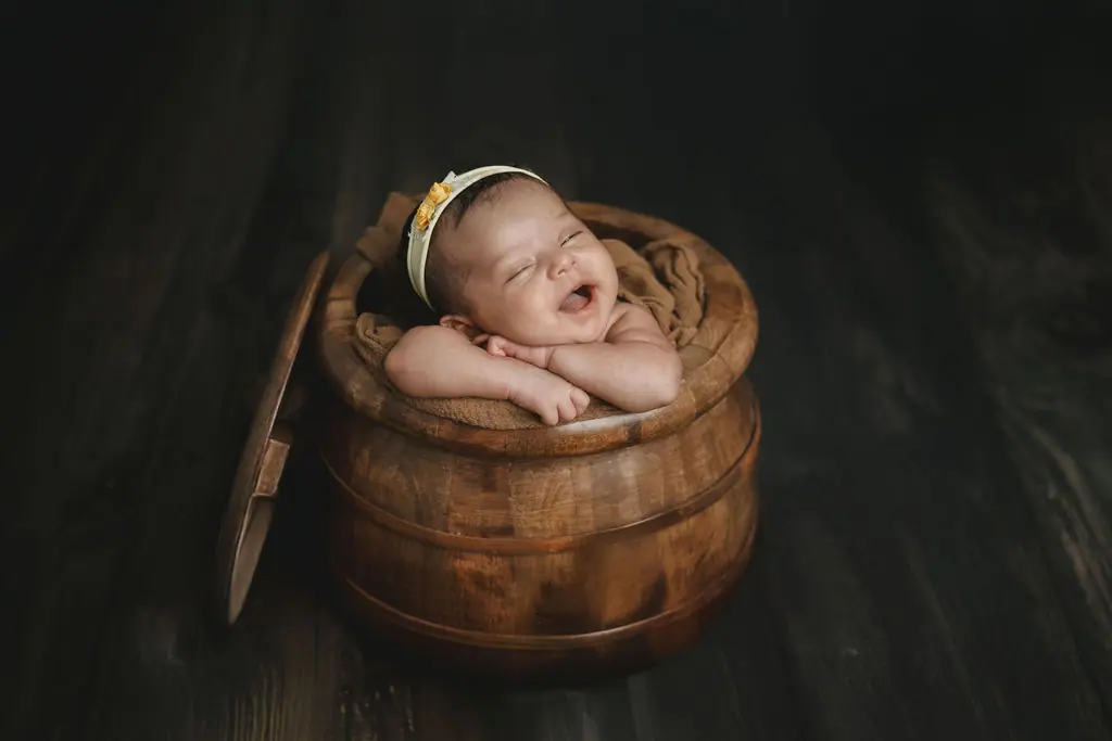 Smiling sleeping newborn with folded arms in a wooden pot, in a cream headband on a dark wood floor, newborn photoshoot at WonderBump Studio, Bengaluru