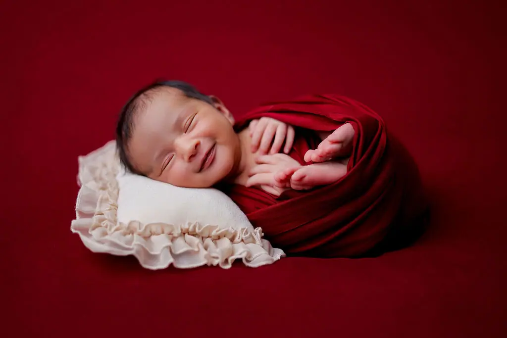 Smiling sleeping newborn swaddled in deep red, resting on a ruffled cream pillow against a red backdrop, newborn photoshoot at WonderBump Studio, Bengaluru
