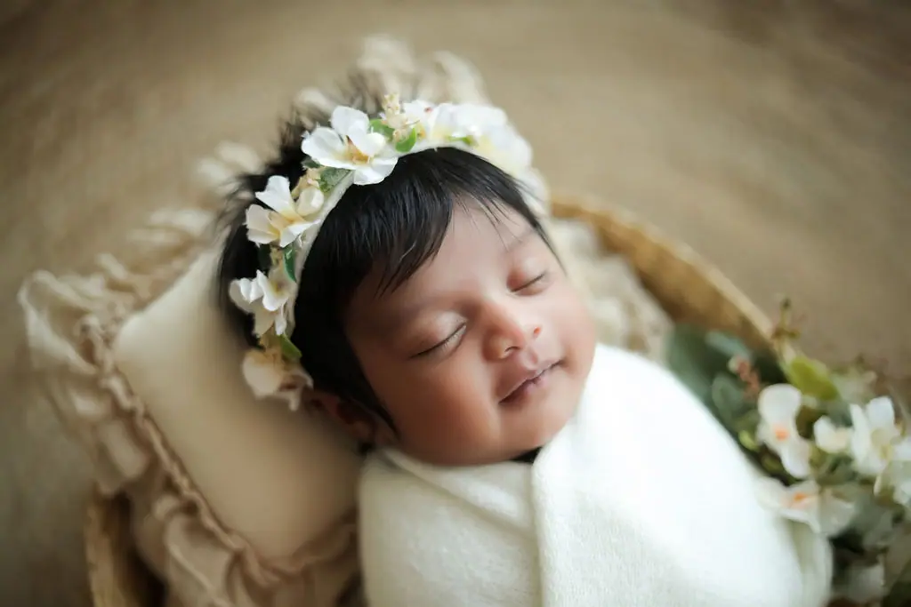 Smiling sleeping newborn in a white flower crown and cream wrap on a ruffled pillow in a basket, newborn photoshoot at WonderBump Studio, Bengaluru
