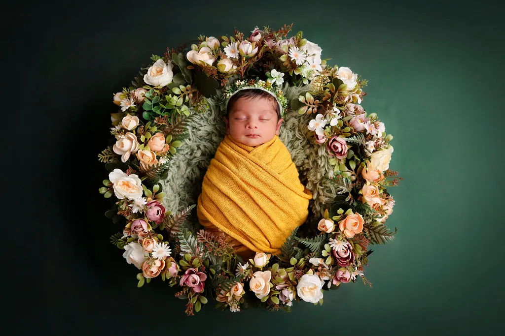 Sleeping newborn swaddled in mustard yellow inside a ring of cream and pink flowers on a green backdrop, newborn photoshoot at WonderBump Studio, Bengaluru