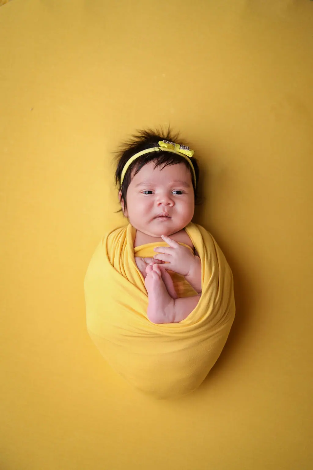 Newborn swaddled in a yellow wrap with a bow headband, awake on a yellow backdrop, newborn photoshoot at WonderBump Studio, Bengaluru