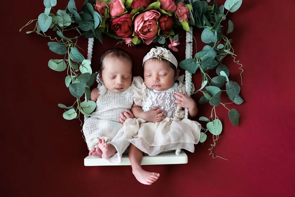 Two sleeping newborns in cream lace on a white swing with roses on deep red, newborn photoshoot at WonderBump Studio, Bengaluru