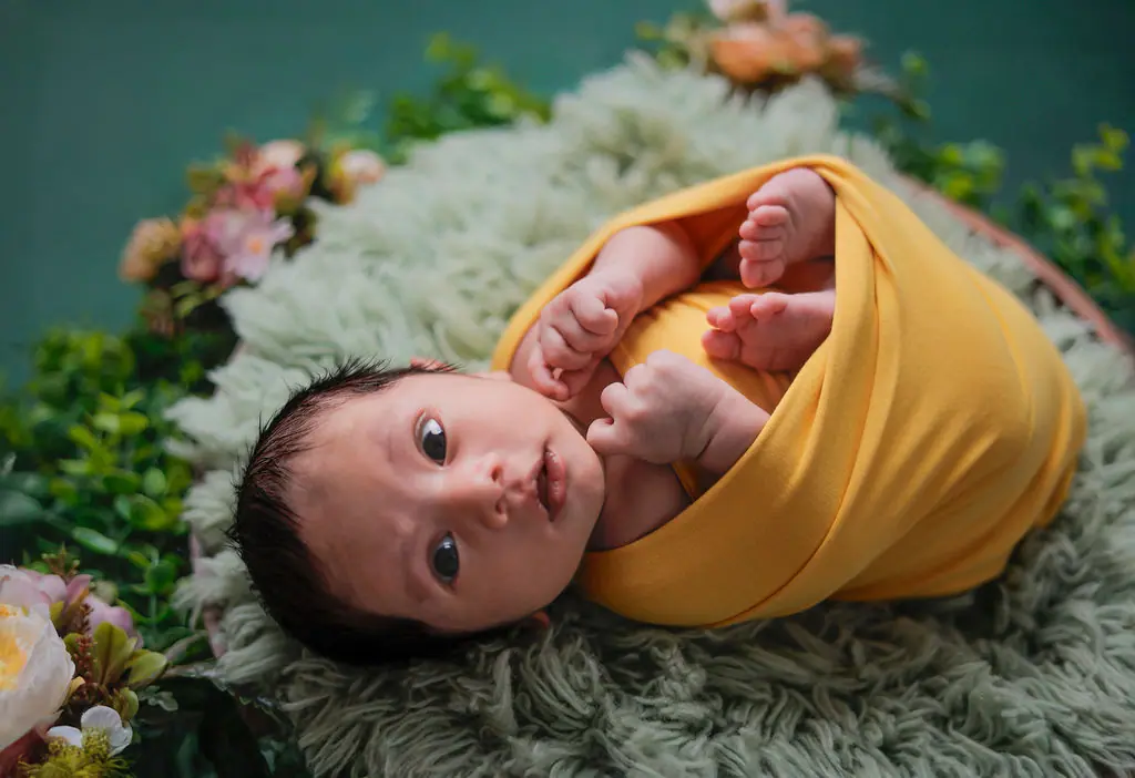 Wide-eyed newborn wrapped in mustard yellow on green faux fur, framed by flowers and greenery, newborn photoshoot at WonderBump Studio, Bengaluru