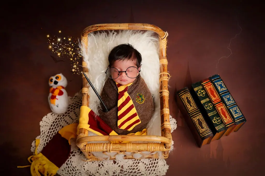 Sleeping newborn in round glasses and a striped tie in a cane basket with a toy owl and books, wizard-themed newborn photoshoot at WonderBump Studio, Bengaluru