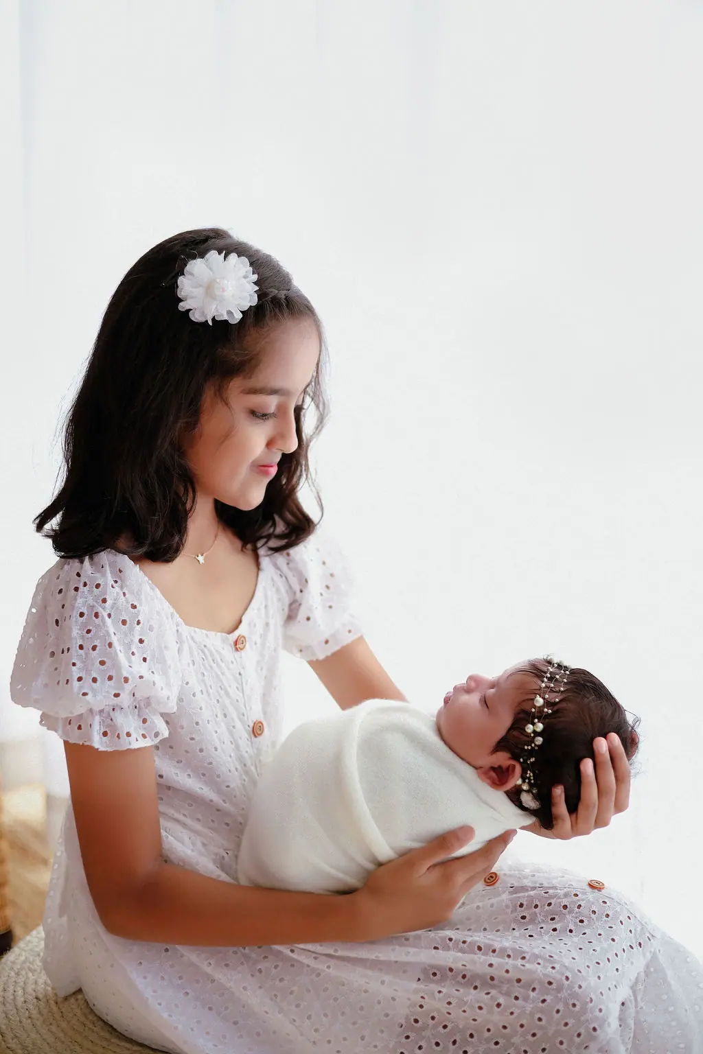 Older sibling in a white eyelet dress cradling a swaddled newborn, sibling newborn photoshoot at WonderBump Studio, Bengaluru