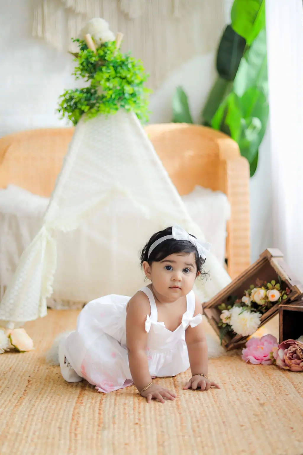 Baby crawling in a white dress and bow headband beside a lace teepee and peonies, toddler photoshoot at WonderBump Studio, Bengaluru